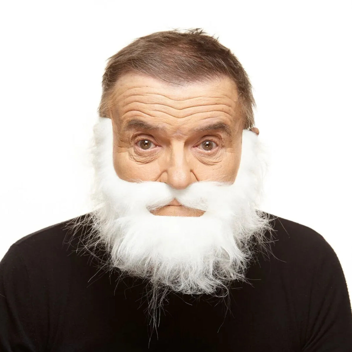 En man med en intensiv, stirrande blick bär ett yvigt och spöklikt vitt lösskägg med en matchande mustasch, vilket ger honom utseendet av en havsgast eller en galen sjöman.