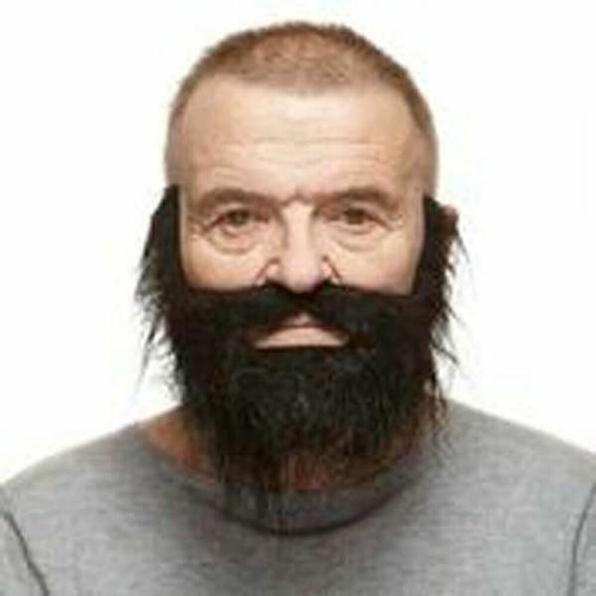 En man bär ett svart, tätt lösskägg med mustasch som täcker kinder, haka och överläpp. Skägget skapar en kraftfull och maskulin look mot en neutral bakgrund.