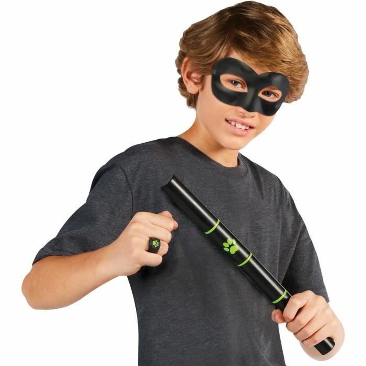 En ung pojke ler och håller i en svart stav med gröna detaljer samtidigt som han bär en svart ögonmask och en ring med ett tassavtryck på fingret.