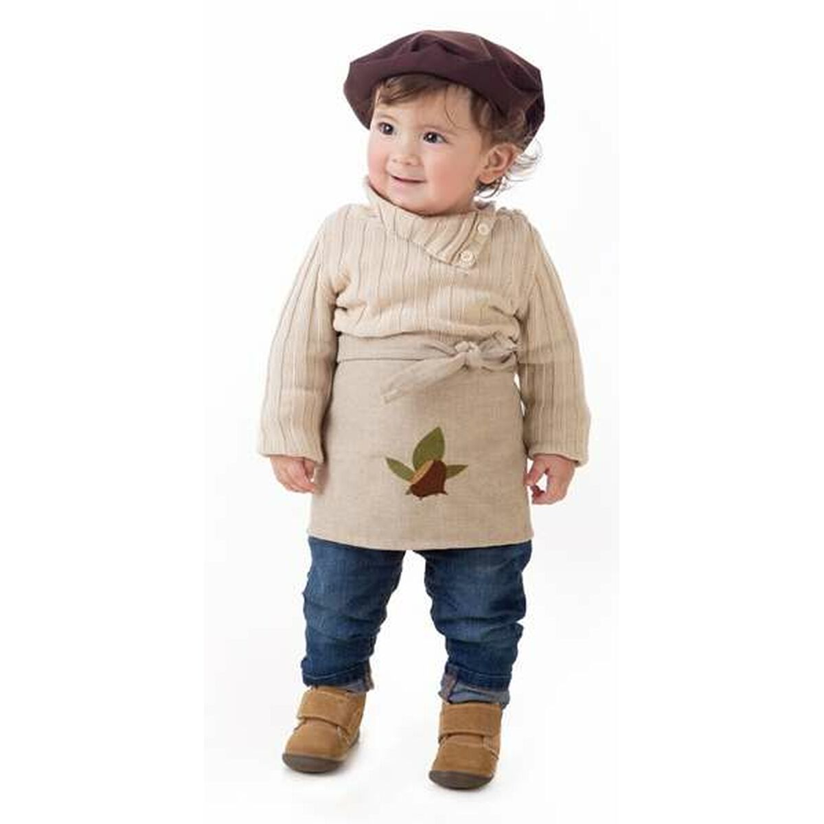 Ett litet barn i en beige tröja, jeans och en brun basker, med en beige förklädesliknande del framtill som är dekorerad med ett kastanjmotiv.