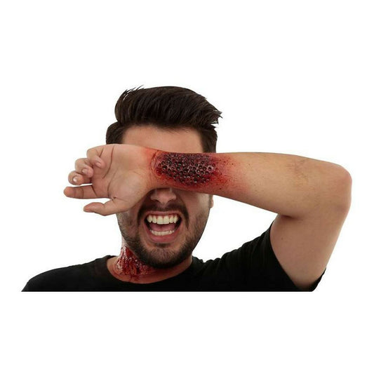 En man med en extremt realistisk latexapplikation som ser ut som ett utbrott med små hål och blod på underarmen, täckande ögat.