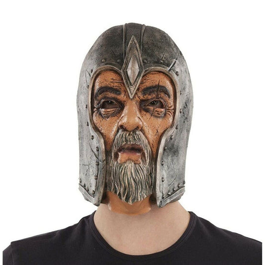 En person bär en heltäckande latexmask som föreställer en gammal, ärrad krigare eller riddare med grått skägg och en sliten, nitad metallhjälm.