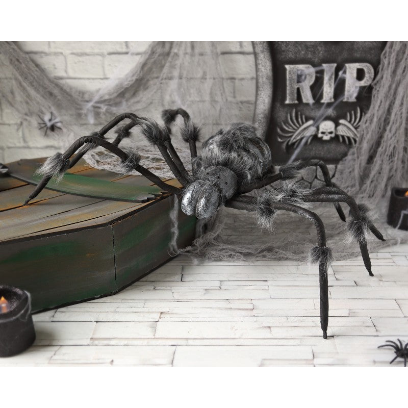 En Gigantisk Benig Spindel Dekoration sitter på en kista i en kuslig Halloween-miljö med spindelnät och en gravsten.