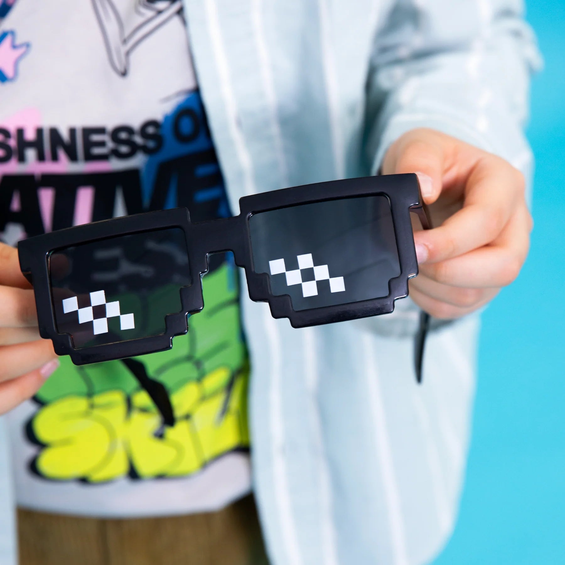 Ett par svarta pixel solglasögon med mörka linser och vita pixelrutor hålls i händerna mot färgglad bakgrund.