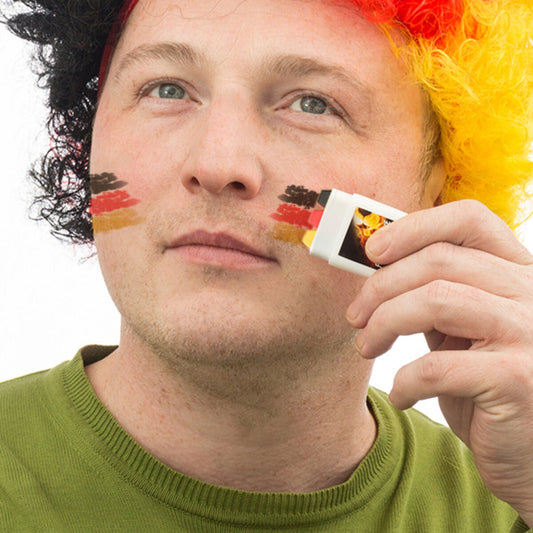 Man med tyskt sminkstift applicerar flaggan på kinden, bär svart-röd-gul peruk.