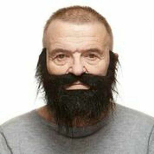 En man bär ett svart, tätt lösskägg med mustasch som täcker kinder, haka och överläpp. Skägget skapar en kraftfull och maskulin look mot en neutral bakgrund.