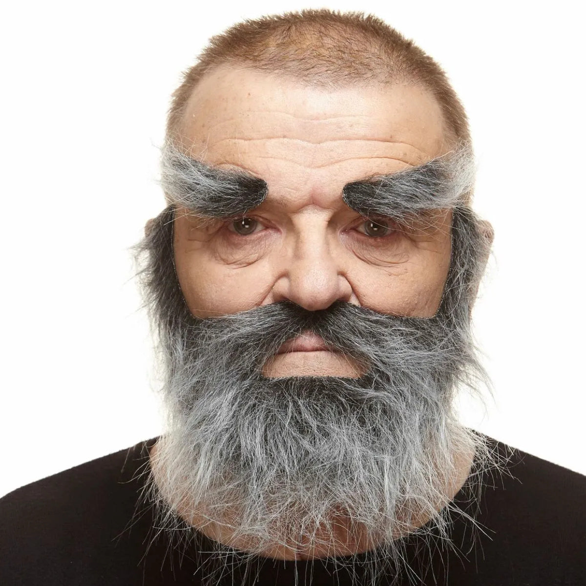 En man med en allvarlig blick bär ett stort, yvigt och vilt lösskägg-set i svart och grått, med en tjock mustasch och extremt buskiga ögonbryn, vilket ger honom utseendet av en gammal, väderbiten pirat.